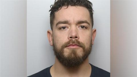 A mugshot of a man with short black hair, a dark beard and a black shirt.