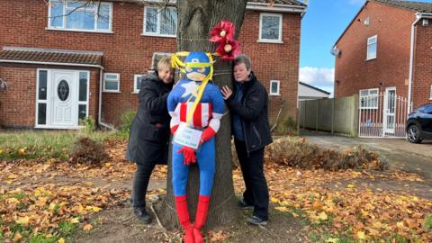 Two women hug a tree on a residential street. They are both wearing black winter coats and black trousers. There is a large super-hero doll tied to the tree.