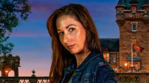A close-up image of Rachel Duffy from County Down. She is looking at the camera with a neutral expression. She has long, dark hair and is wearing a blue denim jacket. Behind her is the traitors Castle, with the sky getting darker as the sun sets.