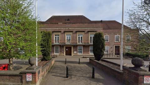 Two-storey brick-built municipal building with white-framed French doors on the upper floor, with a railed balcony across the front. There are white-framed windows on the ground floor, and a dark wooden door in the centre. There are two white flagpoles flanking a paved entrance path. There are trees on either side of the path and a low brick wall.