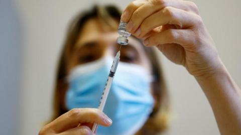 A needle going into a vaccine vial, a woman is holding both of them and is in the background. She is slightly blurred out but has blonde hair and a blue face mask on. 