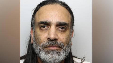 A police mugshot of a man with long dark hair, a grey beard and brown eyes.
