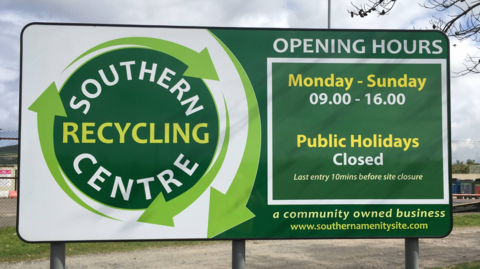 Picure of the recycling sites sign in Port St Mary. It reads it's opening times and logo to the left.