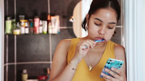 A young woman scrolling her phone as she brushes her teeth, looking hopeless