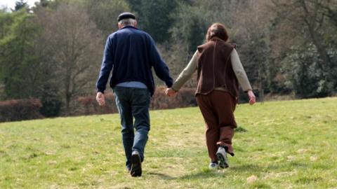 A man in a cap and a woman in a brown coat walk across grass with their backs to the camera