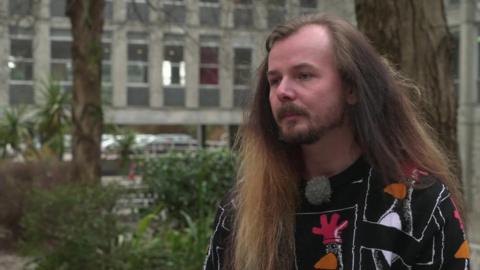 Jack Wilkes is seen outdoors here, next to trees with a grey office-type building behind him. He has long hair, a beard and a moustache, and is wearing a black sweater with pictures of the Wallace and Gromit penguin character Feathers McGraw on it. 