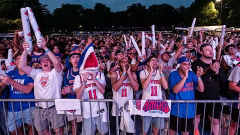 a crowd of fans wearing Knicks gear gather in a park to watch one of the finals games