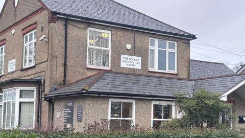 The front of The Hollies Surgery, which is a beige-coloured, two-storey pebble-dash house. A sign reads THE HOLLIES FAMILY DOCTORS SURGERY.