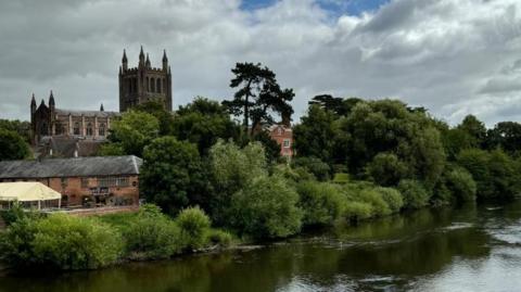 A flowing river with trees and bushes alongside, historical buildings and a cathedral in the background. 