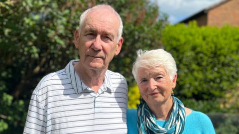 Brian Millin stands on the left with partner Maggie Reeve on the right. Brian is wearing a white polo shirt with black and grey stripes. Maggie is wearing a bright blue top with a blue scarf. 