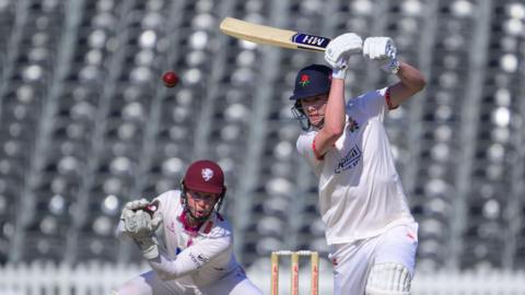 Rocky Flintoff driving a red cricket ball for Lancashire with his bat held high. He is wearing cricket whites and a dark blue Lancashire helmet with the Red Rose insignia on the front.