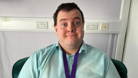 George is sitting in a green chair in front of a wall and some sockets. He is wearing a pale blue shirt and a purple lanyard. He is smiling at the camera.