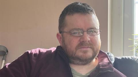 Mark has thin rectangular glasses and is wearing a burgandy fleece unzipped at the collar. He has short stubble and cropped brown hair.