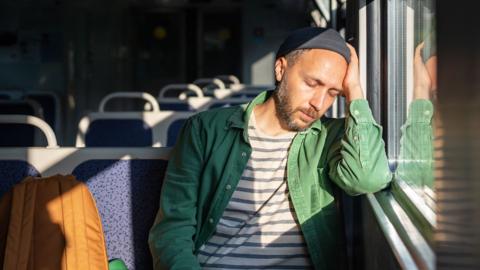 A man wearing a striped t-shirt, open green shirt, and a hat leans on the window ledge of a train while closing his eyes.