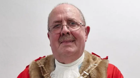 A man in glasses, with short hair and moustache, and wearing chains of office over red ceremonial clothing, looks at the camera.