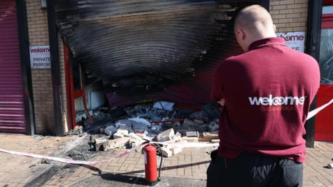 A Welcome Organisation employee wearing a maroon shirt surveys the damage caused to the exterior of the building with a fire extinguisher and bricks laying on the ground