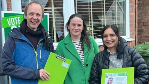 A man on the left (Jonathan Chilvers) smiling in a bluejacket holding a green clipboard next to Clare Bryan who smiles in a green jacket. On her left is Hema YellaPragada who is also smiling holding a green clipboard, she wears a black jacket.