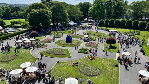 An aerial view of Lister Park in Bradford, a structured garden with flower beds, trees and bushes and lots of people milling about with parasols dotted around. 