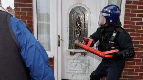 A police officer in riot gear smashes into a front door with a large red tool. 