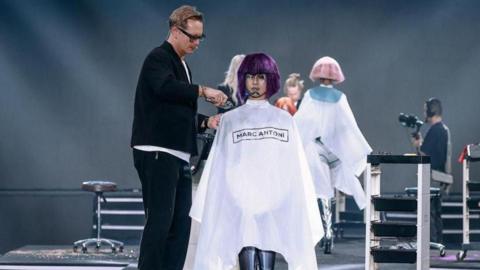 Bruno Marc Giamattei is cutting a purple wig on a mannequin. He has glasses and a black blazer on.