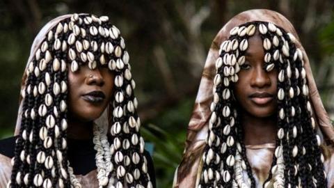 Two women stand near trees. Their clothes are visually striking and their facial expressions are calm. In Dakar, Senegal - Saturday 7 December 2024