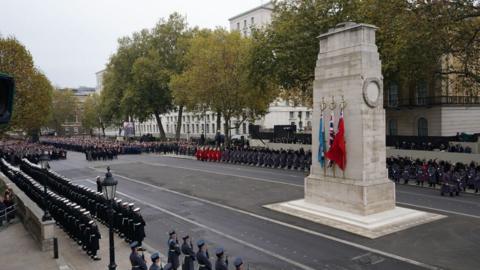 A remembrance ceremony at The Cenotaph in Whitehall, London. Flags of the United Kingdom, Royal Air Force, and Red Ensign are prominently displayed and uniformed military personnel are lined up in a formal formation. Members of the public and officials observing the event from behind barriers.