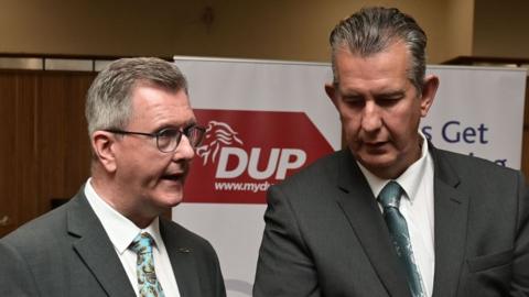 Two men, both with grey hair, wearing grey suits with blue ties. They are stood in front of a DUP banner and engaged in conversation. On the left, Jeffrey Donaldson is wearing black rimmed glasses and appears to be speaking.