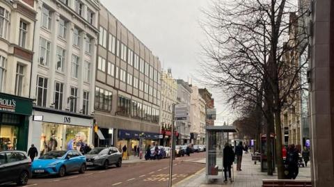 Image of the upper end of Royal Avenue/Donegall Square, there are various people on the pavement on both sides and blue and grey cards to the left.