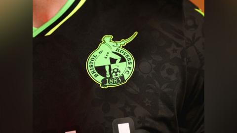 Close-up of the new Bristol Rovers football kit - showing a bright green badge on a black, textured shirt.