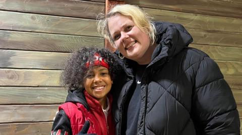 A mum and son wearing coats smile at the camera in front of a wooden wall 