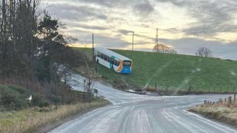 White single-decker bus with blue, green and yellow markings, in a green grassy field, having gone off the road on what appears to be an icy bend.