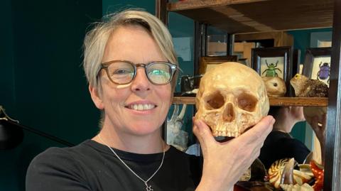 A woman holding what looks like - but definitely is not - a skull, in the background is a cabinet of beetles, shells and a brain