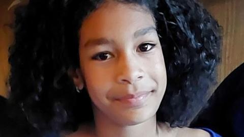 Candid photo of Kaliyah Coa, who died after falling into the Thames. She is 11 and is wearing a blue top and smiling at the camera with big brown eyes and curly hair.