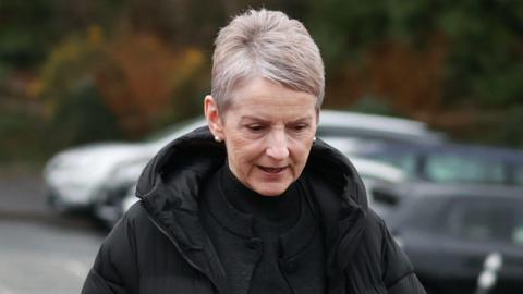 Lady Eleanor Donaldson, with short grey hair, wearing a black top and a black coat with a hood. There are parked cars in the background.