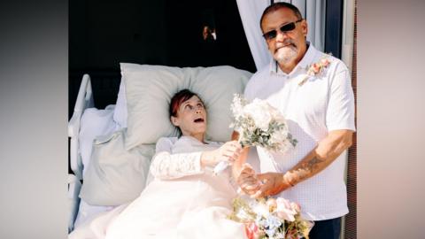 A woman in a hospital bed is pictured next to a man in a short sleeved shirt after their wedding. The woman is holding a bouquet of white flowers and is wearing a wedding dress