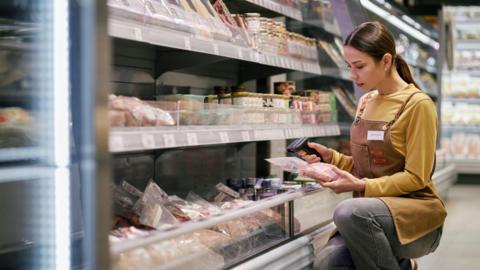 A stock image of a shop worker kneeling in front of a fridge in a supermarket scanning a product. She has brown hair worn in a ponytail.