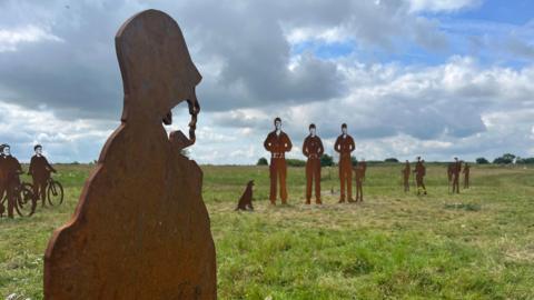 Steel statues of silhouettes on Salisbury Plain. Three soldier statues can be seen in the distance. A statue of a person wearing a hat can be seen in the foreground of the image. Other smaller statues can be seen in the distance of people riding bikes and walking with hiking sticks. 