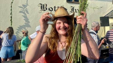 Lucy Dermody, wearing a straw hat, holding a bunch of plucked nettle stems, outside the thatched Bottle Inn pub