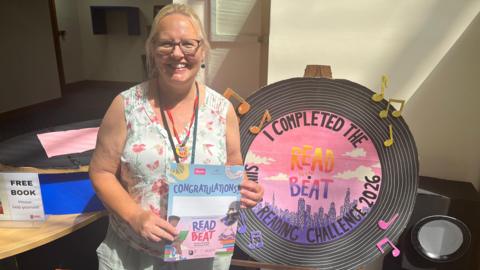 Senior Librarian Julie O'Grady is standing indoors holding a “Read to the Beat” certificate. Beside her is a large display resembling a vinyl record, which says on it “I completed the Read to the Beat Reading Challenge 2026,” decorated with musical notes.