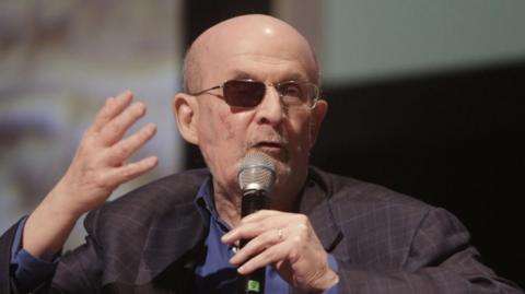 Salman Rushdie speaking into a microphone, he is wearing glasses where one eye section is entirely black so you cannot see what is behind it.