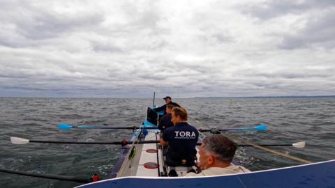 Four people in a rowing boar at sea, with shore visible on the horizon.