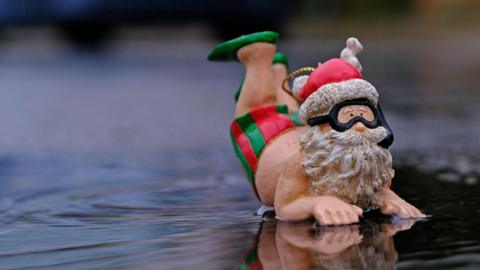 A Father Christmas doll with red hat, but otherwise naked except for red and green swimming trunks and green swimming shoes. He appears to be clinging on to the edge of a pool of water.