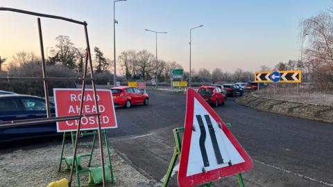 A picture of a roundabout with cars on it, stuck in a queue. There are also road signs to indicate that a nearby road is closed.