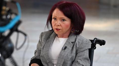 Pam Duncan-Glancy seated in a wheelchair wearing a checked blazer and white top, with a camera positioned in the foreground.