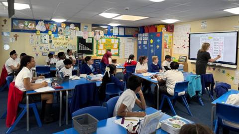 A classroom with children in white shirts or red jumpers sitting around blue desks and a woman is writing on a whiteboard.