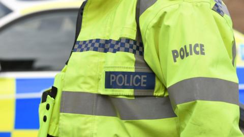 A police officer wearing a high-vis police jacket stands in front of a blue and yellow chequered police vehicle. Only the officer's torso is visible. 