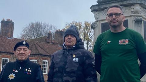Three men stand in front of a war memorial, wearing military medals on civilian clothes and looking seriously at the camera. The man on the left wears a black beret, black gilet with a blue jumper underneath and dark trousers. The man in the middle wears a grey beanie hat and puffer coat with camouflage detail and blue cargo trousers. The man on the right wears a green T-shirt with a black long-sleeved top underneath and blue jeans. 