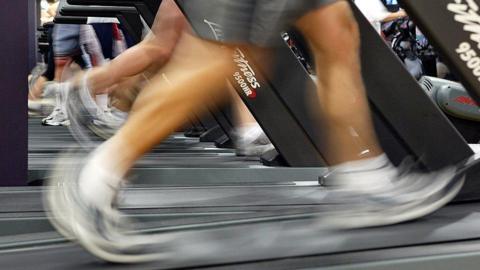 A person's feet can be seen as they run on a treadmill inside a gym. They wear white and black trainers with grey shorts. Other people running on treadmills can be seen behind them.