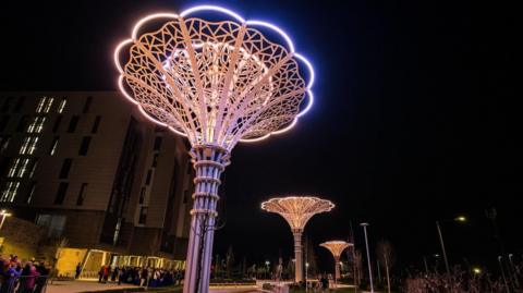 Three towering metal structures have ornate filigree in the shape of roses and are lit up with white lights in the night sky