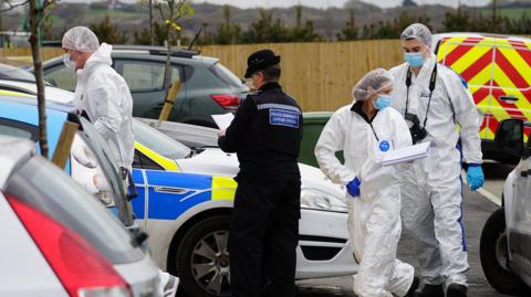 Three people wearing white suits entering a property, while a police community support officer looks at a piece of paper.